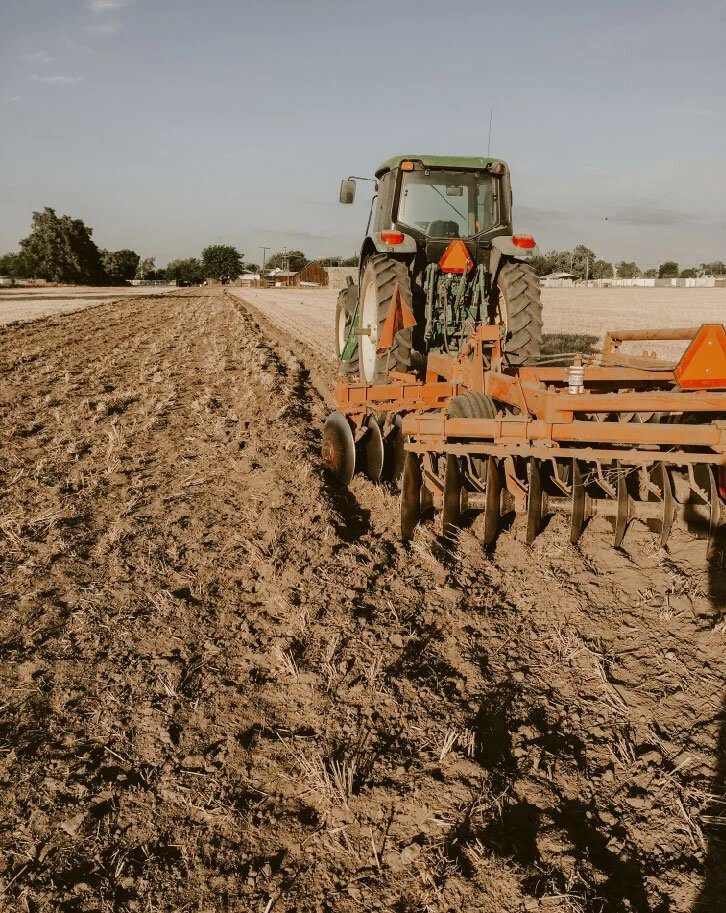 Green tractor pulling an orange disc harrow across a field of dark, freshly turned soil